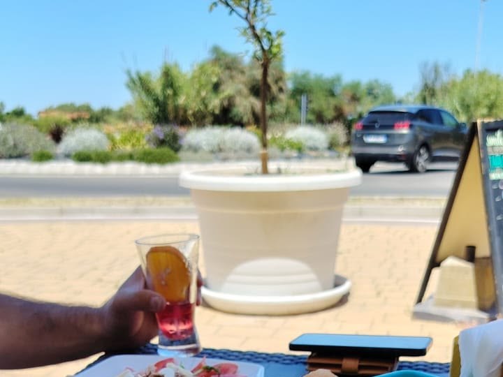 Mano con bicchiere di drink rosso e fetta d'arancia su tavolo all'aperto, vaso bianco e auto grigia sulla strada