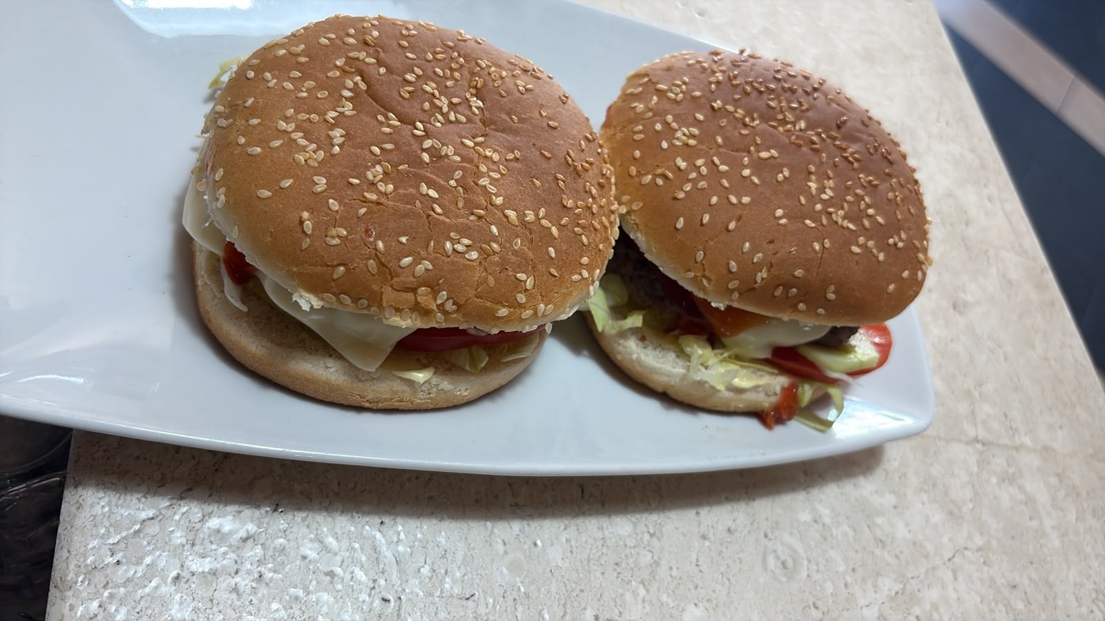 Due hamburger con panino al sesamo, formaggio, lattuga e pomodoro su un piatto bianco rettangolare, piano in marmo chiaro
