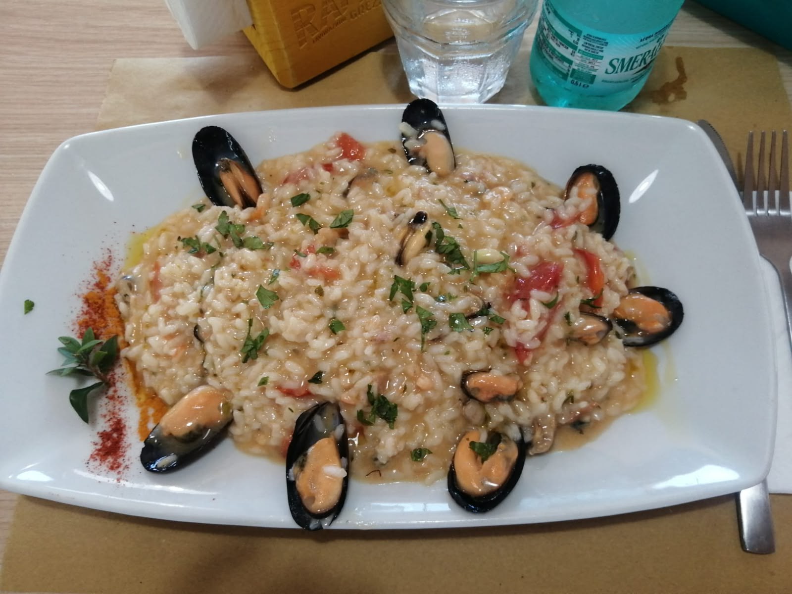 Risotto con cozze, pomodorini e prezzemolo su piatto bianco rettangolare, con paprika, origano e bottiglia d'acqua Smeraldina